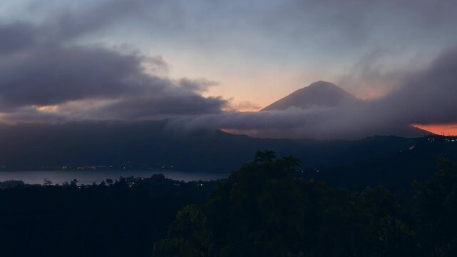 Majestic Mount Abang Rising Above Lake Batur During Sunrise, Dramatic Clouds Flow Across Mountain Slopes Above Forested Valley, Serene Wilderness And Volcanic Landscape