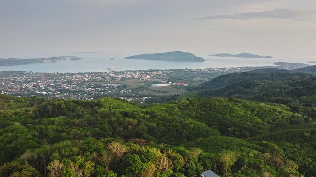 Phuket island's tropical landscape with lush green hills and a thriving coastal city landscape bordering the tranquil bay, dotted with islands and boats under a cloudy sky. Aerial view drone flight
