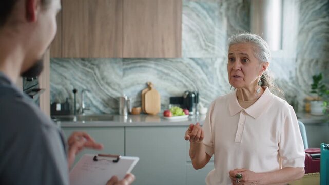 Elderly woman describing her household repair needs to handyman writing note on clipboard during residential consultation in kitchen. Over the shoulder view