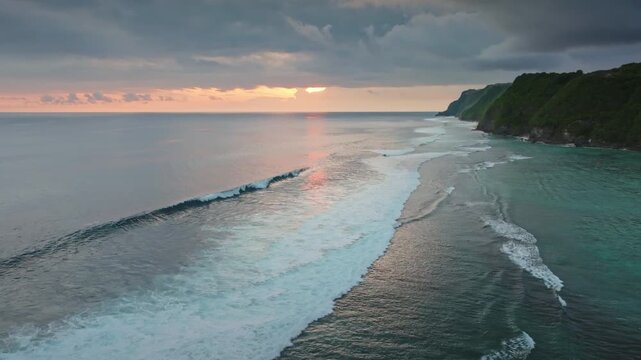 Scenic Bukit Peninsula coastline at sunset where dramatic cliffs meet the vast Indian Ocean, with breaking waves, warm tropical light and tranquil, secluded seascape. Aerial view drone flight panorama