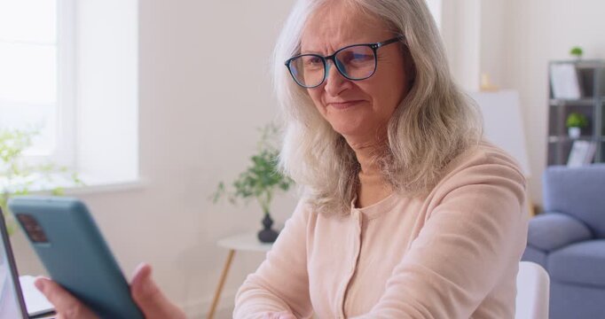 Senior woman in eyeglasses sending text messages on smartphone, adjusting camera before making video call, using modern technologies, sitting at home against home interior. Old people