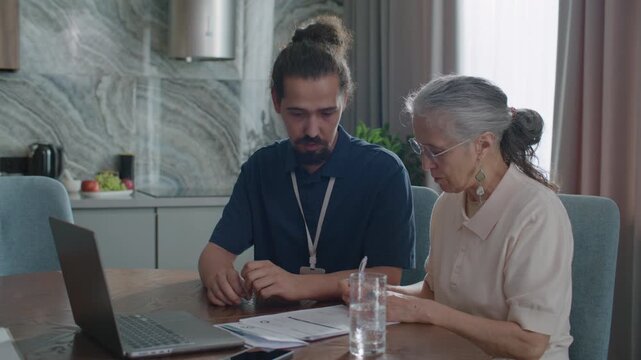 Young male social worker with ID badge helping senior woman to review and sign utility bills or financial documents at home, both sitting at table in living room with laptop on it