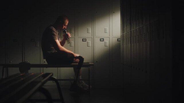Man with Prosthetic Leg Sitting in Locker Room, Hydrating with Bottle while Focused on Smartphone, Watching Training Tutorials or Sports Content. Motivation, and Lifestyle of Modern Adaptive Athlete.