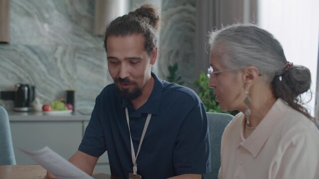 Young male social worker explaining financial document details to cheerful senior woman, she smiling and asking him questions during home care visit or consultation
