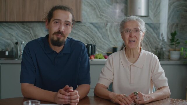 Senior woman and her adult grandson sitting together at kitchen table, looking at camera and speaking during remote video call or online consultation. POV shot