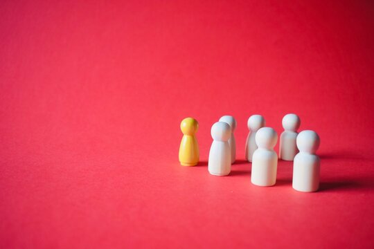 A single yellow wooden peg doll leading a group of white peg dolls on a vibrant red background, conceptualizing leadership, individuality, and team management.