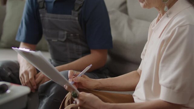 Close up view of senior woman talking to plumber or repairman and signing service contract on clipboard while sitting on sofa in sunlit living room