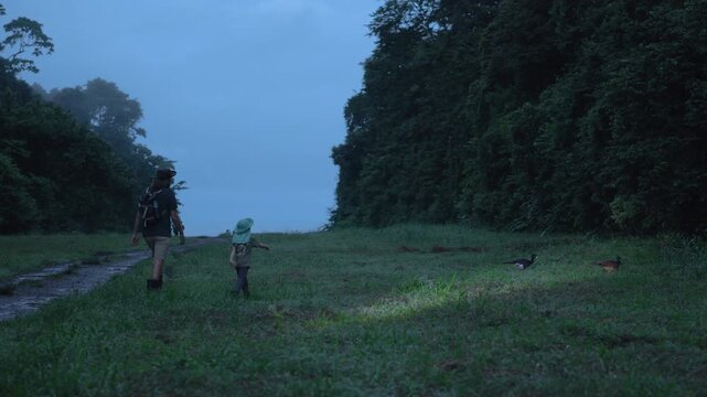 Father and son walk at night with flashlights along grassy path in dense tropical rainforest. Adult and child wear hiking gear and rubber boots while wild birds forage nearby in misty dusk atmosphere.
