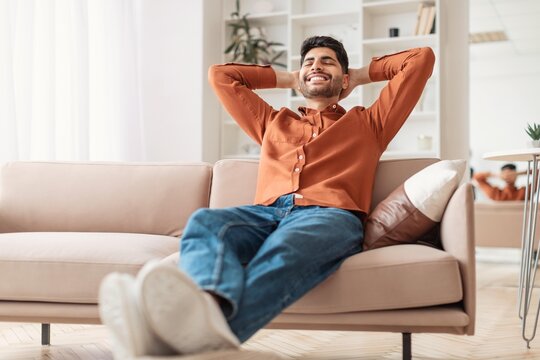 Smiling Young Hispanic Man Sitting On Comfortable Couch With Feet Up, Enjoying Relaxation At Home In A Cozy Living Room. Reflecting Positive Vibes And Leisure Lifestyle Concept.