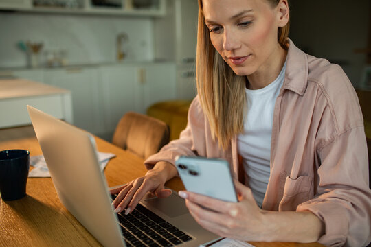 Young woman working from home with laptop and smartphone in kitchen