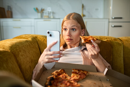 Young woman eating pizza and using smartphone on couch at home