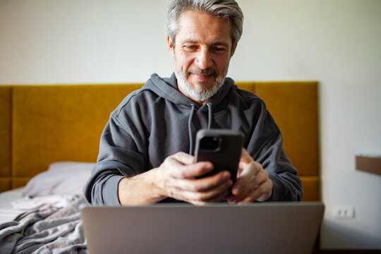 Mature man using smartphone and laptop in bedroom at home