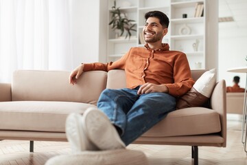 Obraz premium Smiling Young Man Sitting Comfortably On Sofa In Modern Living Room, Enjoying Leisure Time And Relaxing After A Long Day, Creating A Cozy Home Vibe. Lifestyle Concept.