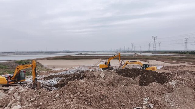 Two yellow excavators operating on a construction site beside wetlands under overcast sky