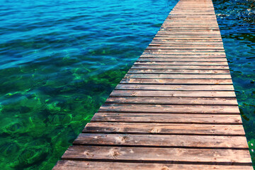 Fototapeta premium Weathered wooden dock extends diagonally over strikingly clear water, blending vivid turquoise and emerald green hues. Sunlight filters through the shallow depths, revealing rocky lakebed below
