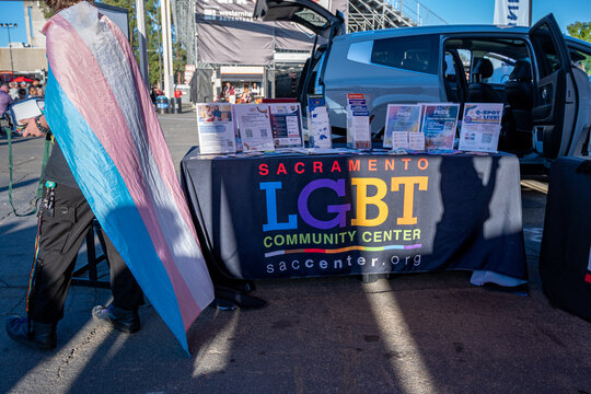 An informational resource booth for the Sacramento LGBT Community Center with an attendee wearing a transgender pride flag cape during a Pride Month event.