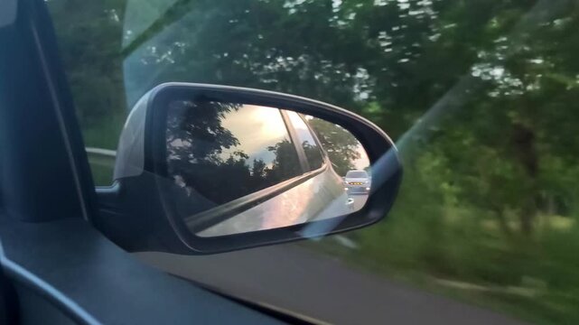 Close-up view of a car side mirror reflecting the road, another vehicle, and a sunset sky while driving past a green roadside landscape.