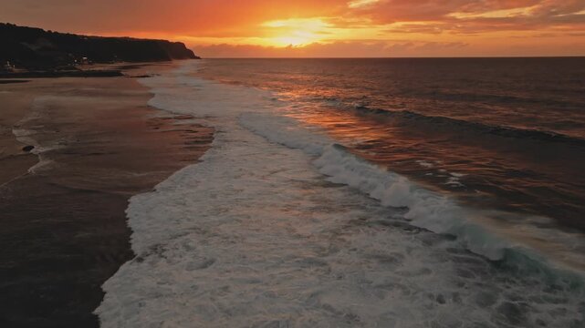 Ocean waves crashing on Melasti Beach shore during a vibrant orange sunset in Bali, Indonesia, highlighting the beautiful coastal landscape and the powerful nature. Drone flight zoom out