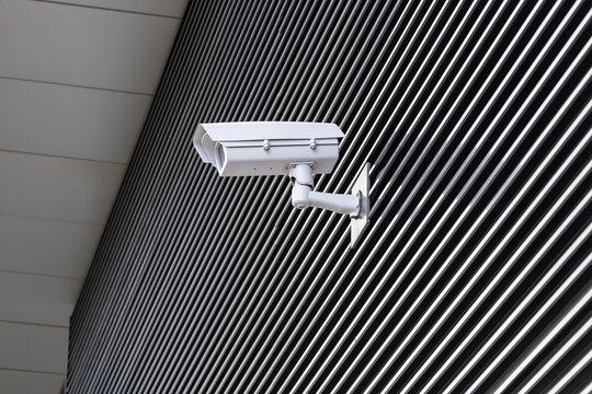 An outdoor CCTV security camera is mounted on a bracket against a modern building exterior featuring striped metal cladding facade. Concept of security, technology, and smart city.