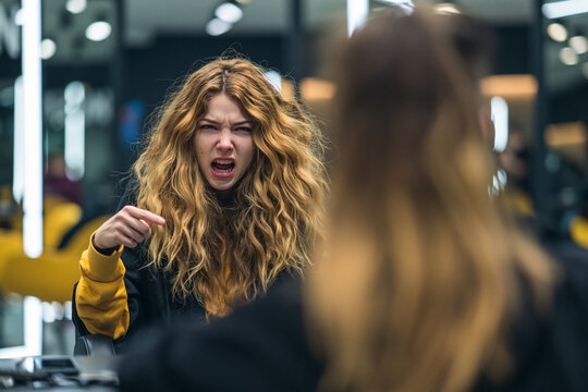 > Over the shoulder view of a furious female customer pointing and yelling at a hairdresser. Dissatisfied client complaining about bad haircut and hair color in a beauty salon. Poor customer service 