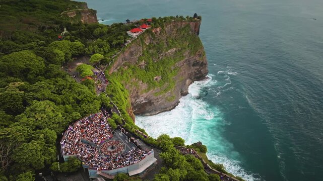 Uluwatu Temple, a sacred Balinese Hindu sea temple. Dramatic cliff overlooking the crashing Indian Ocean, bustling amphitheater with tourists people watching a traditional Kecak dance. Aerial shot