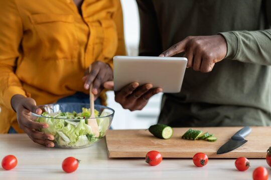 Focused African American Couple Preparing Fresh Healthy Salad Together In Modern Kitchen, Using Tablet For Recipe Guidance. Engaging In Cooking While Enjoying Their Time Together. Health Concept.