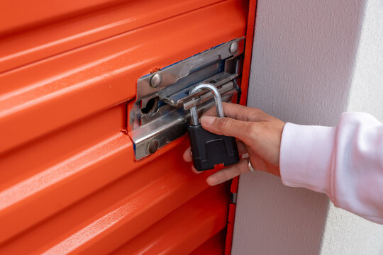 A hand in a white sleeve unlocks a black padlock on a metal latch attached to an orange corrugated storage door, indicating entry to a secured storage unit