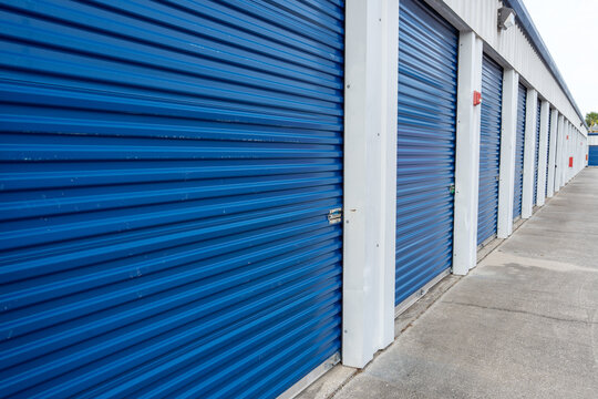 A row of storage units with blue corrugated metal doors and white frames, aligned along a paved corridor that leads into the distance, emphasizing an organized and structured facility