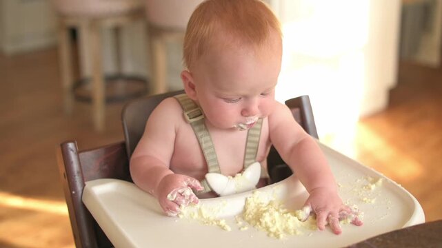 Caucasian baby in high chair engages with food, moving from tasting to creating a mess, with a bright kitchen backdrop featuring wooden flooring and warm, soft lighting enhancing the scene