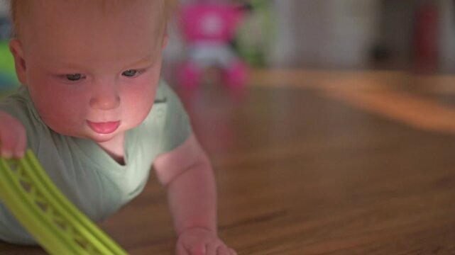 Young child with light red hair stretches out to grasp a green toy on a wooden floor, illustrating a moment of exploration in a bright indoor play area with colorful objects in the background