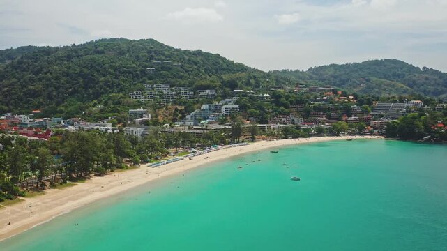 Phuket tropical coastline showing Patong beach, turquoise waters, sandy shores, lush green hills and a vibrant resort town. Summer vacation, travel to Thailand. Aerial view drone flight