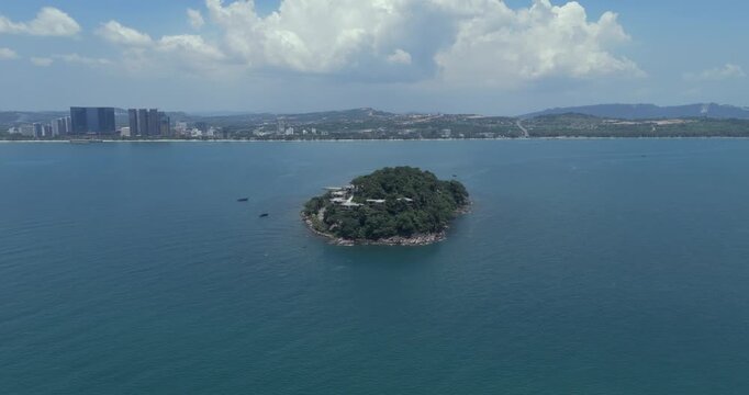 Aerial turning view of a lush island with modern villas tucked into the forested hillside, set in a vast, tranquil blue sea.