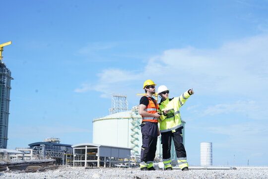 Male Caucasian engineers and their male foreman colleagues, all in uniform and helmets, are discussing and planning inspections of maintenance work while standing at an oil storage tank 