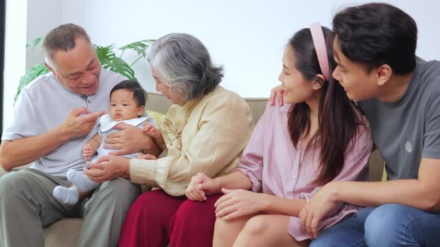 Joyful Asian family members bonding at home. Beautiful moment of grandparents and parents looking at the sweet baby boy with love, laughing and sitting closely on a couch