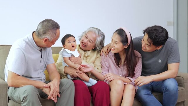 Joyful Asian family members bonding at home. Beautiful moment of grandparents and parents looking at the sweet baby boy with love, laughing and sitting closely on a couch.