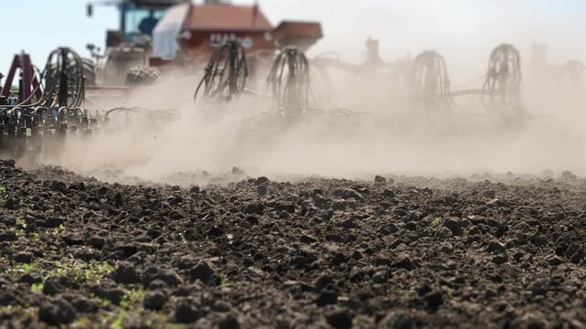 Modern seeding machine operating across cultivated farmland and raising dust during planting season. Agricultural equipment performing large scale field preparation and crop production.