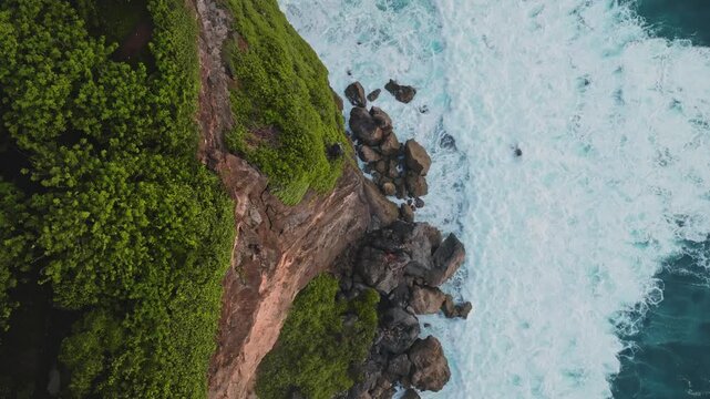 Dramatic cliff separating lush green vegetation from the powerful turquoise ocean, with white foam of crashing waves breaking on the rocky Bali shore. Natural background. Aerial view static shot
