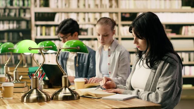 Portrait of smiling students and adult tutor sit together in library, diverse study group at wooden table with books, notebooks and green lamps for collaborative study and learning.