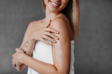 Fototapeta premium Smiling Young Caucasian Woman Applying Moisturizer On Her Arm While Enjoying Skincare Routine At Home, As Part Of Daily Self-care And Beauty Regimen In Relaxed Indoor Setting. Skincare Concept.