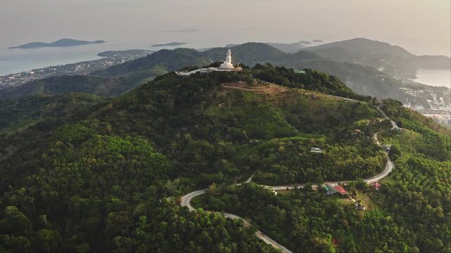 Iconic Big Buddha statue on top of lush green mountain, with a winding road leading up and the coastal towns and Andaman Sea stretching along the horizon. Travel to Thailand. Aerial view drone flight