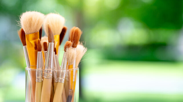 Set of various artist paintbrush in glass holder against a blurry green background of garden. Paint tool for creativity art workshop