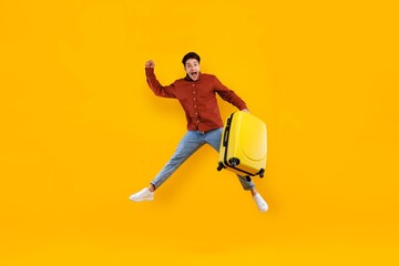 Fototapeta premium Smiling Young Man Of Asian Descent Jumping With Yellow Luggage, Showing Excitement For Travel In Studio Setting. Perfect For Travel Concept, Adventure, Vacation, Excitement.