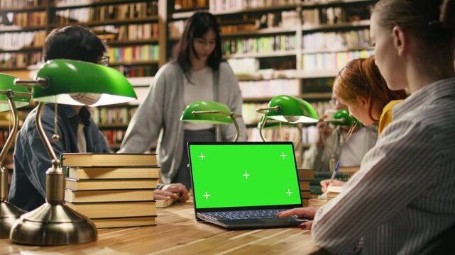 Green screen mockup on laptop inside university library desk, study space surrounded by blurred students and green lamps, Editable screen on device with touchpad, with group study and assignment.