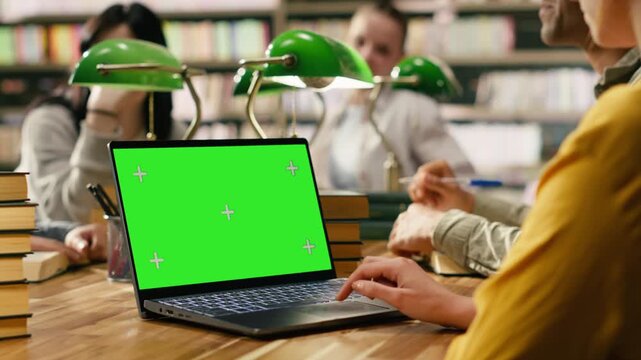 Green screen mockup on laptop with peers at wooden library desk, student hand uses the touchpad near editable screen.