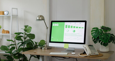 Fototapeta premium Office desk with monitor showing an esg dashboard of environmental, social and governance charts and graphs, surrounded by green potted plants in a professional workspace