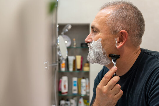 Handsome white-haired beared man shaving off his beard looking in a mirror. Concept of changing style