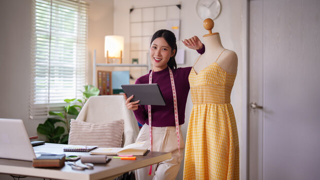 Young Asian female fashion designer presenting yellow gingham dress on mannequin while holding tablet in bright modern home studio workspace with measuring tape