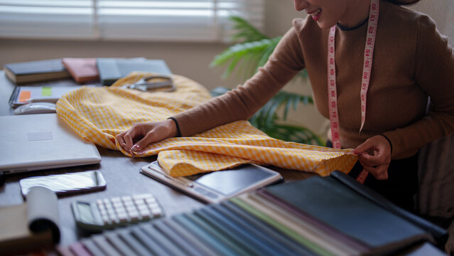 Asian fashion designer examining yellow striped fabric with measuring tape around neck while working at desk with color swatches in bright studio workspace