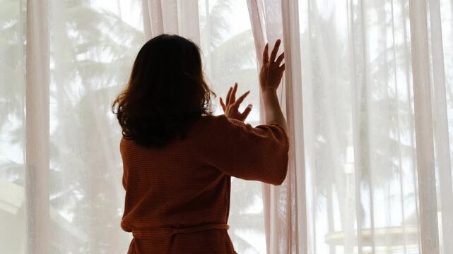 The woman was at the window in the bedroom. She opened the curtains on the window. In the morning and she looks at the view of mountains and trees at sunrise.	