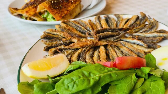 Traditional Turkish fried hamsi anchovies served on a plate with fresh lemon slice and green salad arugula. Delicious local crispy seafood fish dish with appetizing street food wraps on background.